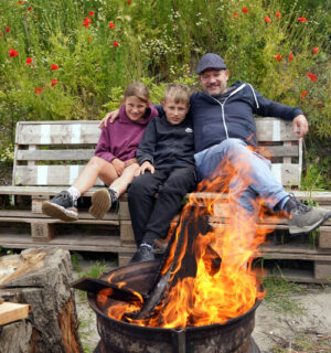 Vader met 2 kinderen op bankje buiten bij het vuur. Op de achtergrond groen met veldbloemen.