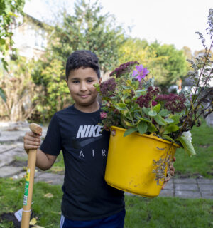 Een jonge, glimlachende jongen staat in de buurttuin en houdt een schep en gele emmer, gevuld met bloemen en planten, vast.