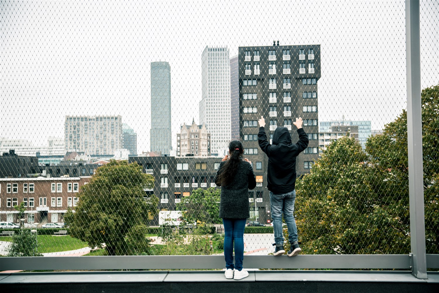 2 kinderen kijken door een gaas naar hoge gebouwen in de verte.